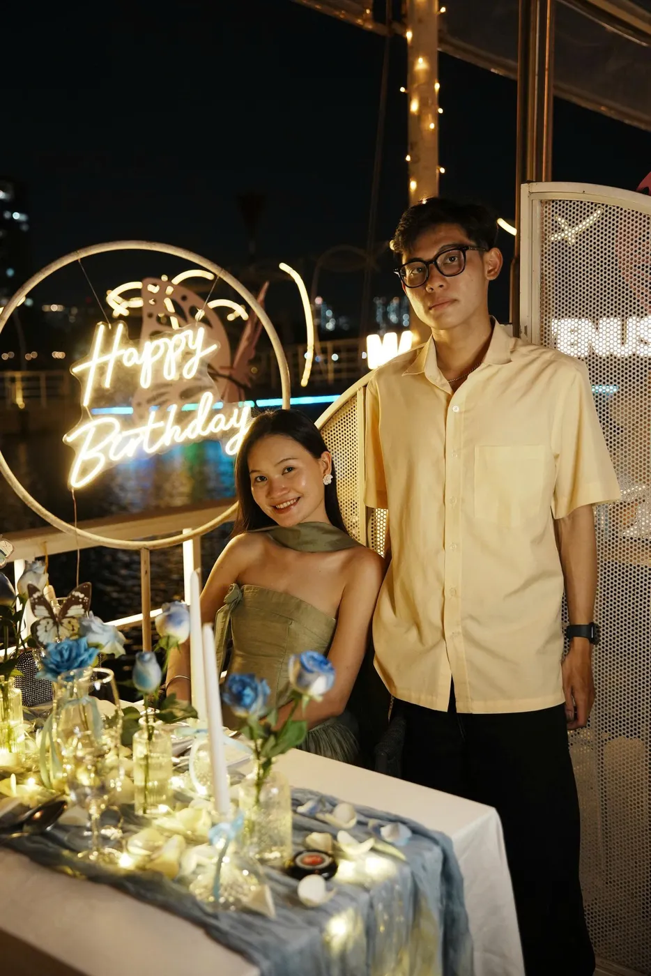 Một cặp đôi cụng ly bên bàn tiệc trang trí hoa hồng xanh, phía sau là biển hiệu neon Happy Birthday và tòa nhà thành phố về đêm