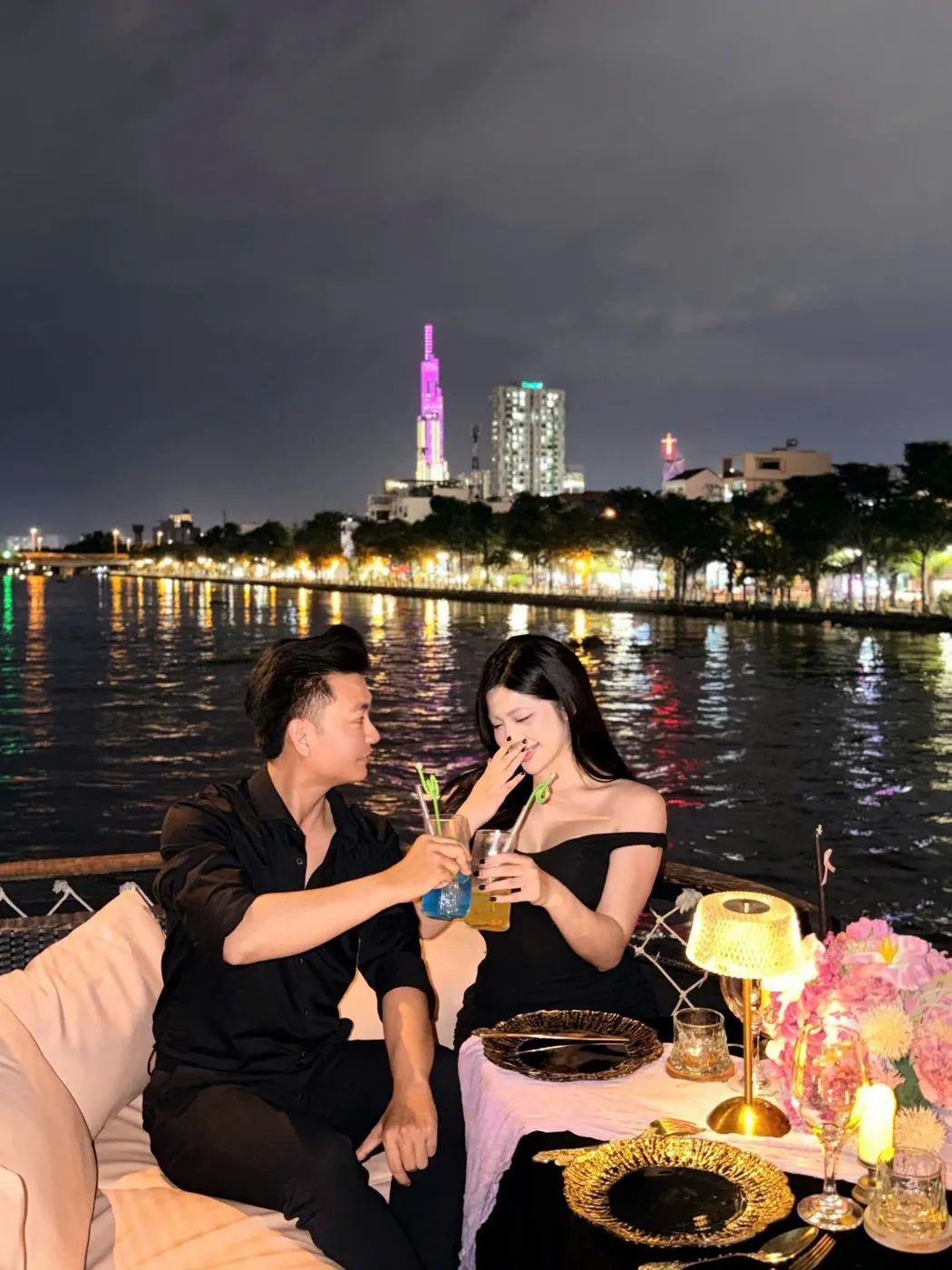 Một cặp đôi ngồi trên du thuyền, cùng nhâm nhi đồ uống với khung cảnh sông Sài Gòn và tòa nhà Landmark 81 rực rỡ ánh đèn phía sau