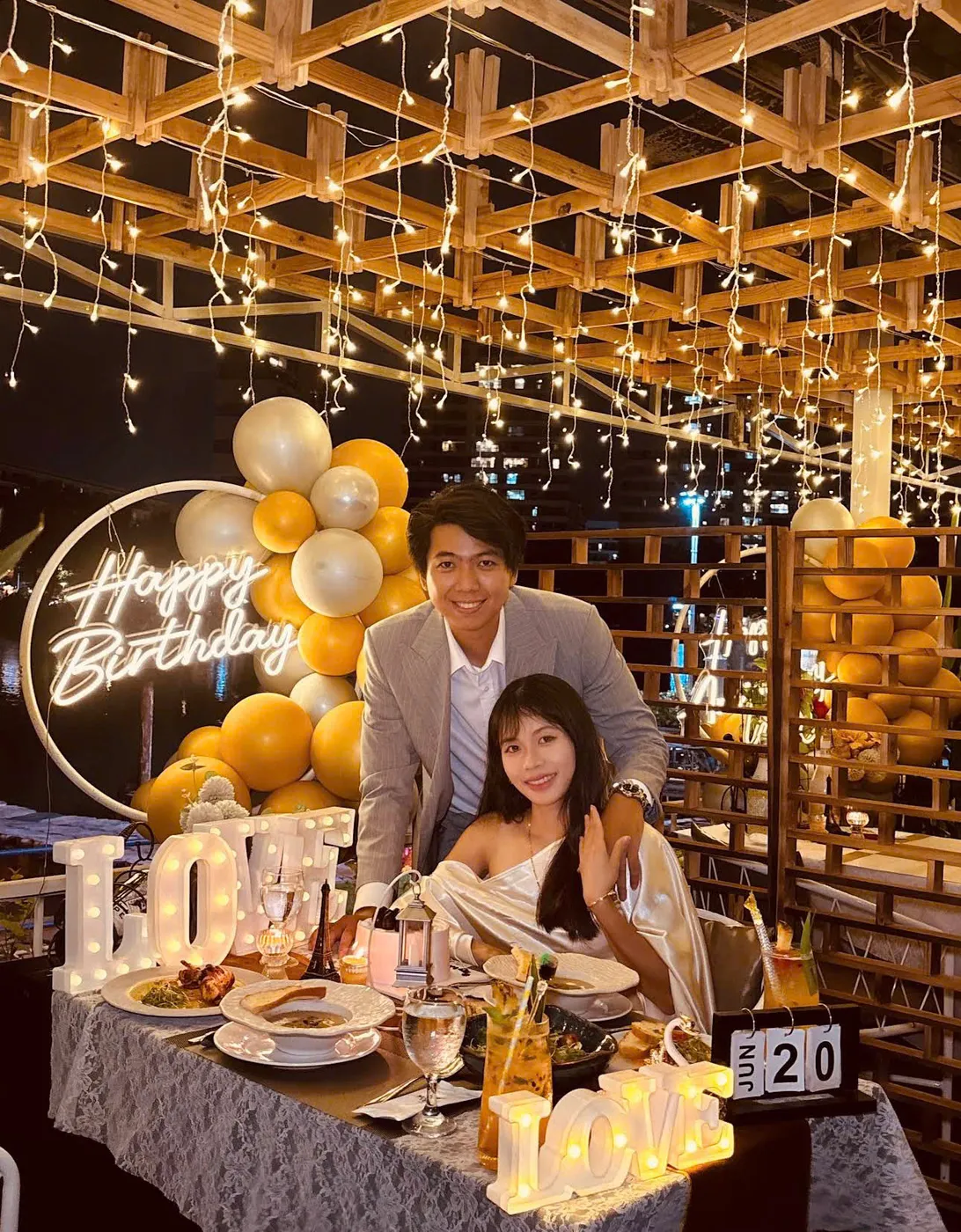 Một cặp đôi chụp ảnh cạnh biển hiệu neon Happy Birthday cùng bóng bay vàng và chữ LOVE phát sáng trên bàn tiệc