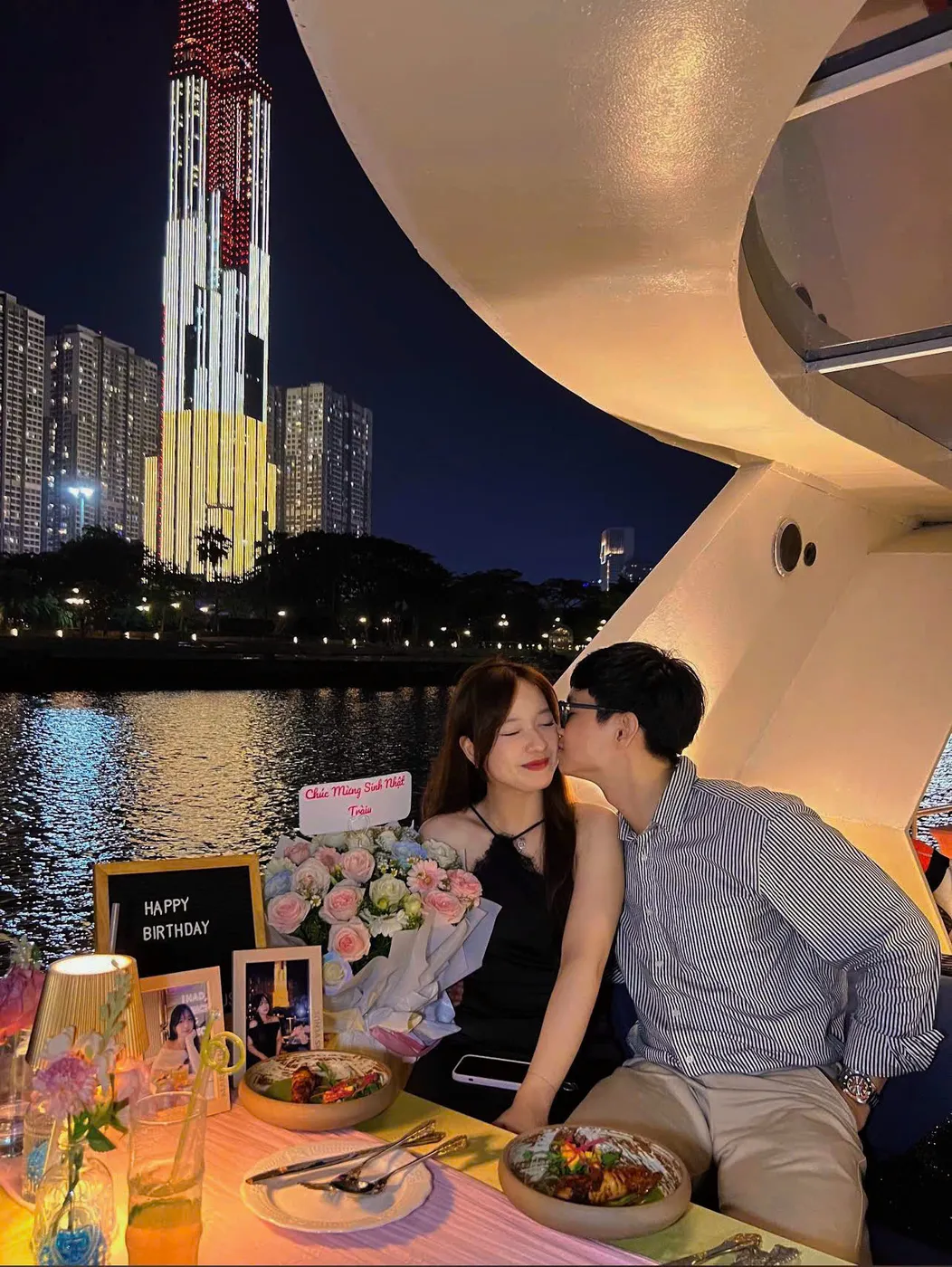 Một chàng trai hôn lên má bạn gái trên du thuyền, phía sau là tòa nhà cao tầng rực sáng và bó hoa mừng sinh nhật trên bàn