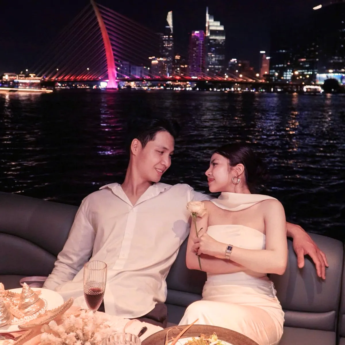 Một cặp đôi tựa vai nhau trên du thuyền trong trang phục trắng, phía xa là cây cầu ánh đèn đỏ và đường chân trời thành phố về đêm