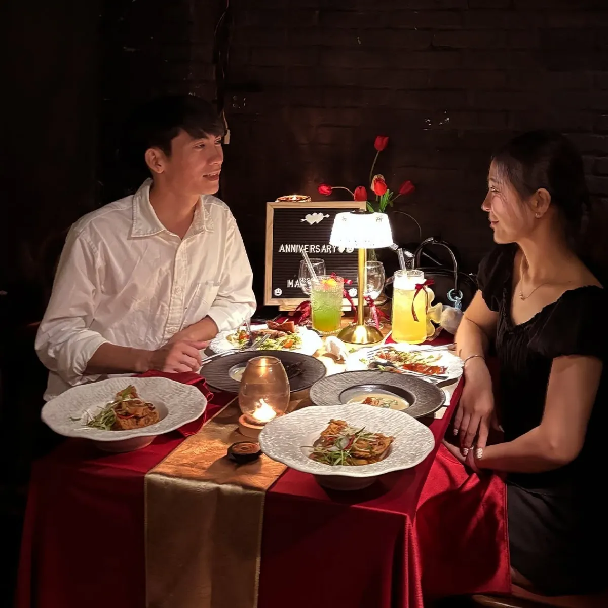 Một cặp đôi trò chuyện bên bàn tiệc kỷ niệm với khăn trải bàn đỏ, nến lung linh và bảng chữ Anniversary trong không gian ấm cúng